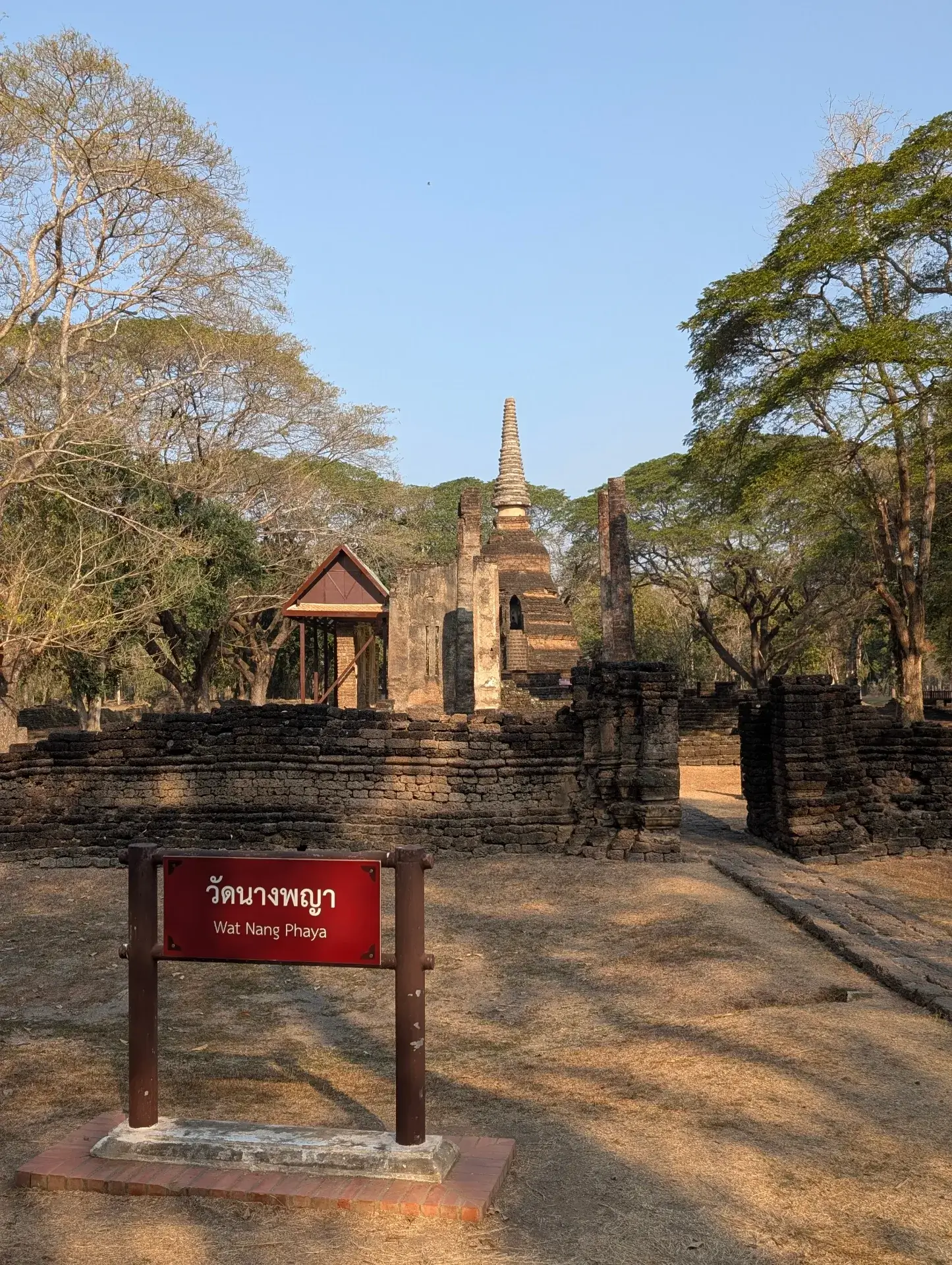 タイのシーサッチャナーライ歴史公園内にある「ワット・ナーンパヤー」。手前に「Wat Nang Phaya」と書かれた赤い看板があり、背景には美しい彫刻が残る仏塔（チェディ）と礼拝堂の遺構が、乾いた大地と木々に囲まれて佇んでいる。
