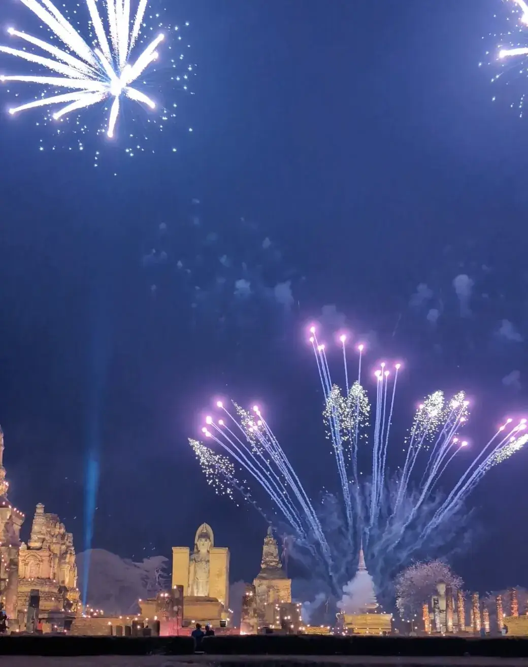 夜のスコータイ歴史公園にて、ライトアップされた仏像や遺跡の真上に打ち上がった大きな花火。白とピンクの光が夜空に広がり、サーチライトと共に祝祭の盛り上がりを象徴する、旅のフィナーレにふさわしい光景。