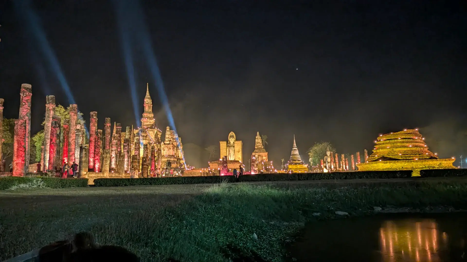 夜のスコータイ歴史公園、ワット・マハータートの広大な全景。数本の青白いサーチライトが夜空へ伸び、赤や黄金にライトアップされた無数の仏塔や柱、大仏が立ち並ぶ。手前の水面にはその光が反射し、幻想的なシンメトリーを形作っている。