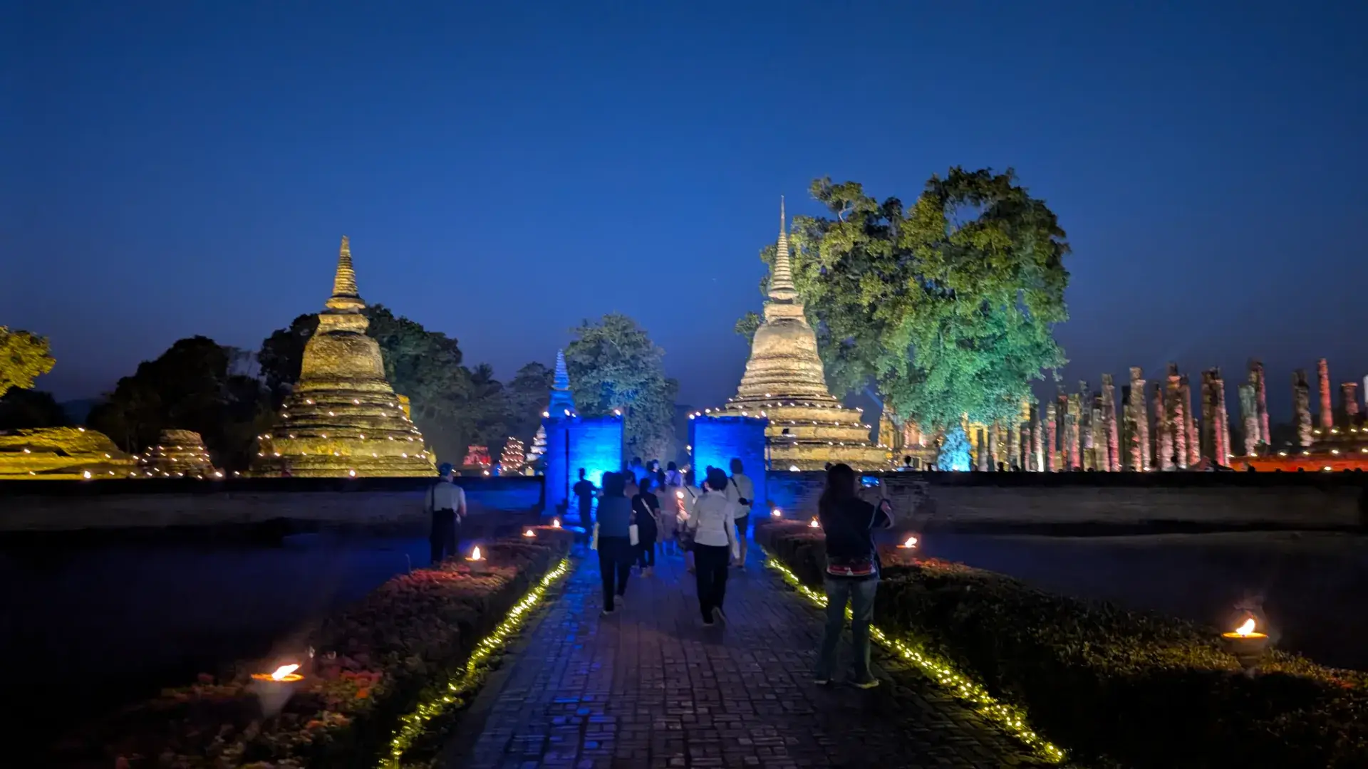 夜のスコータイ歴史公園。黄金色にライトアップされた巨大な仏塔（チェディ）と、青い光に照らされた門。参道の両脇には小さな灯火が並び、多くの観光客がその幻想的な風景の中を歩いている。