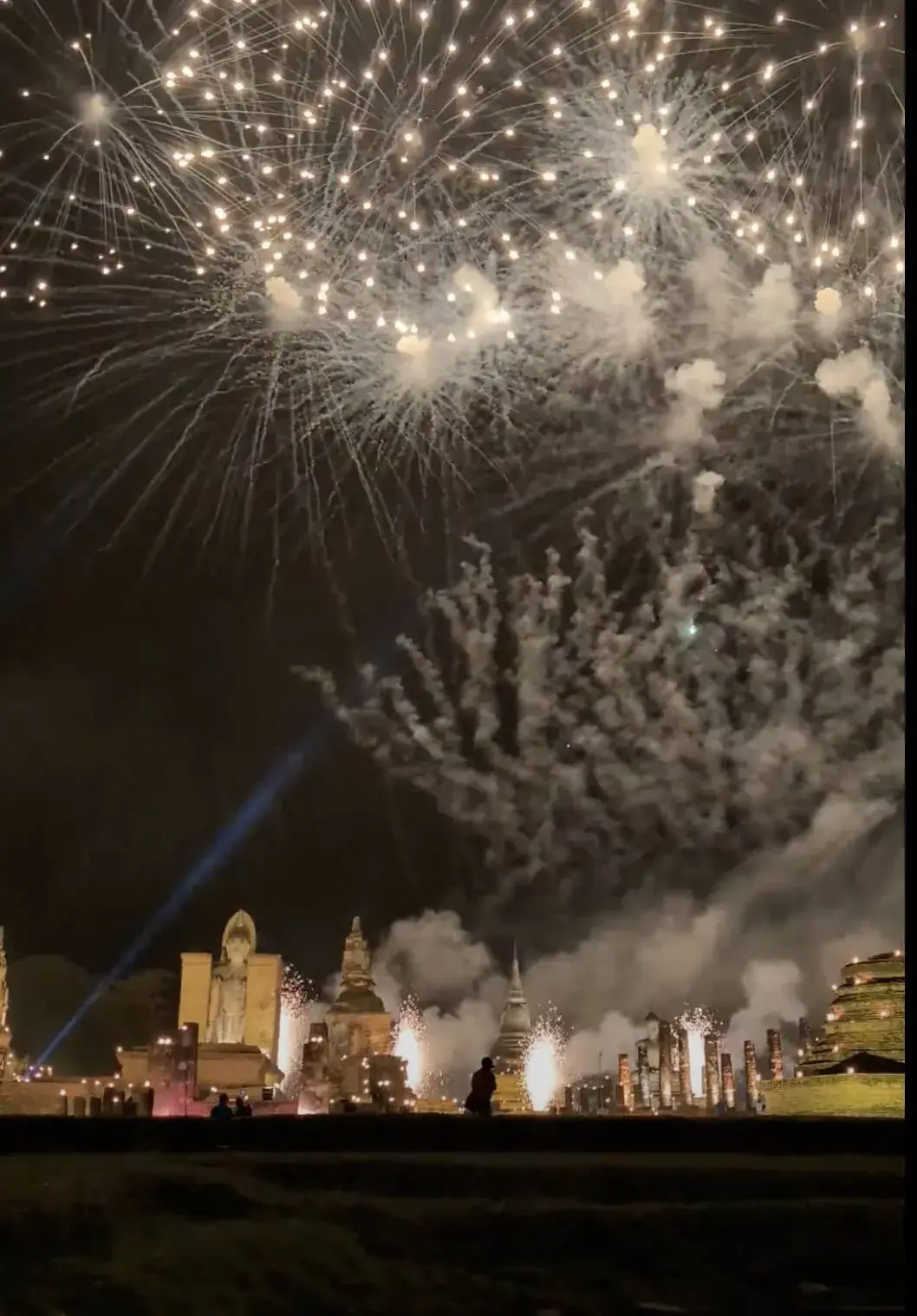 ライトアップされたスコータイ歴史公園の遺跡群の真上に、白、エメラルドグリーン、そして黄金色の巨大な花火が次々と打ち上がる様子。サーチライトの光と花火の煙が混ざり合い、幻想的で力強い祝祭の夜を演出している。