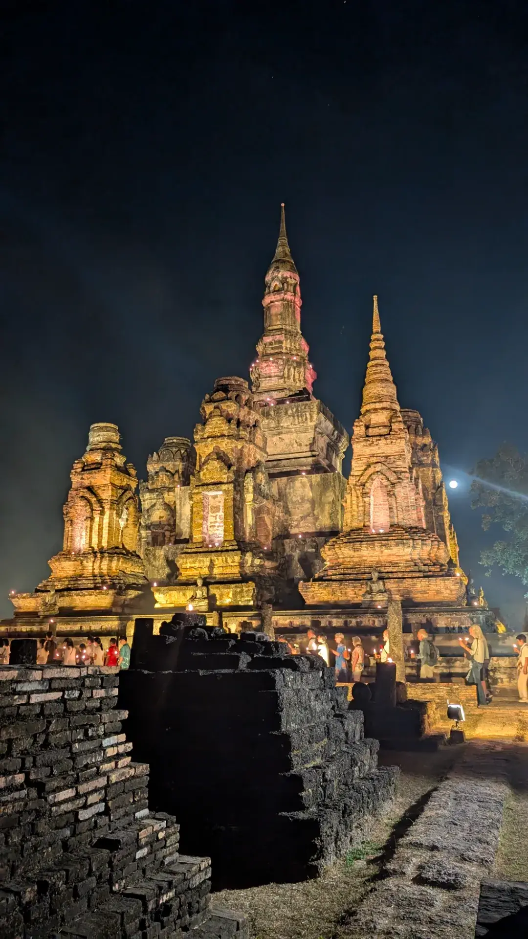 夜のスコータイ歴史公園。黄金色にライトアップされたワット・マハータートの主塔を背景に、手に灯火を持った多くの参拝客が列をなして歩いている。歴史的遺構の重厚な質感と、現代の祈りの光が融合した神聖な光景。