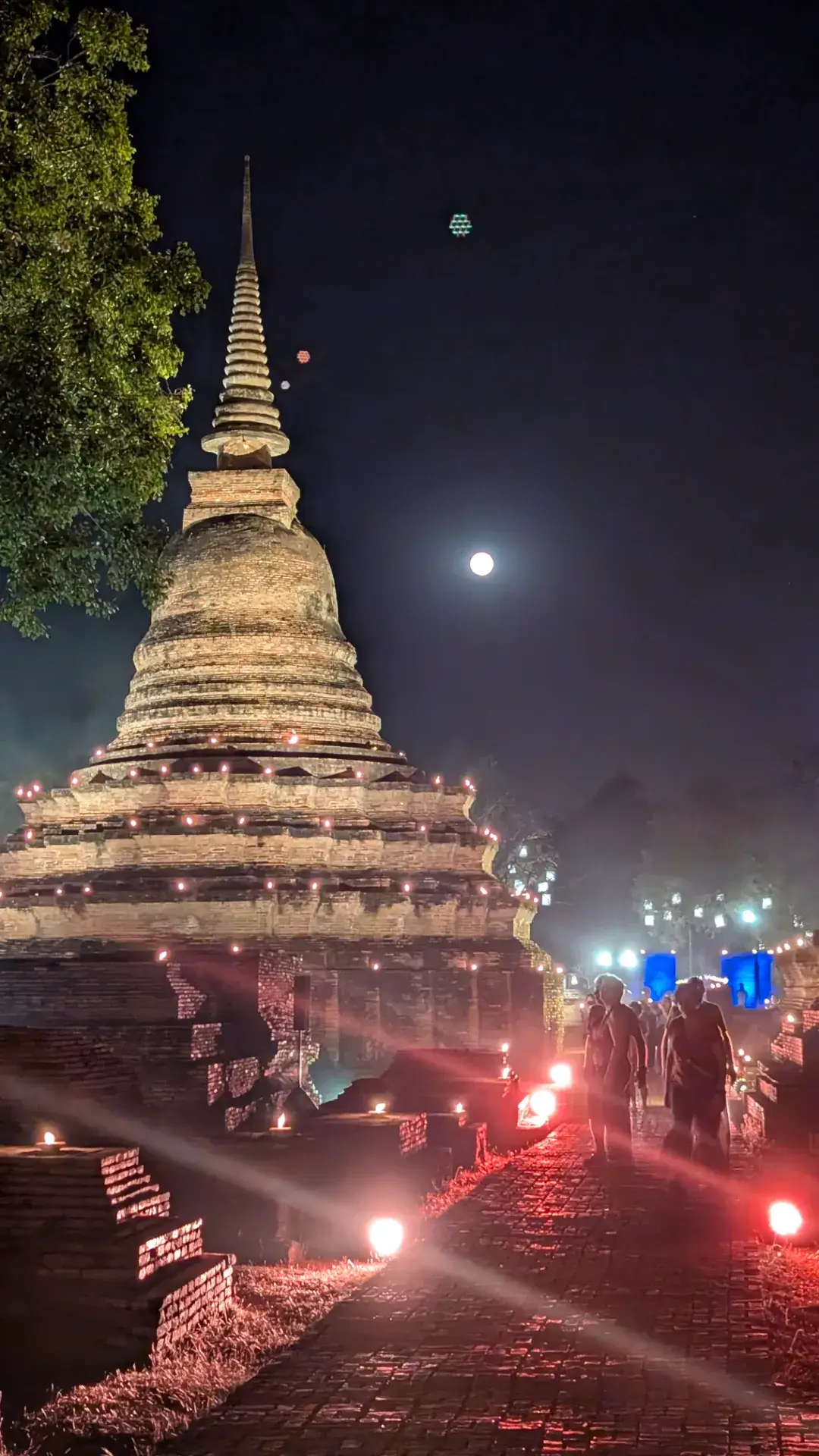 夜のスコータイ歴史公園にて、満月が空に輝く中、無数の小さなオイルランプ（灯明）で縁取られたワット・マハータートの大仏塔。赤みを帯びたライトアップが参道を照らし、シルエットとなった参拝客が歩いている幻想的な光景。