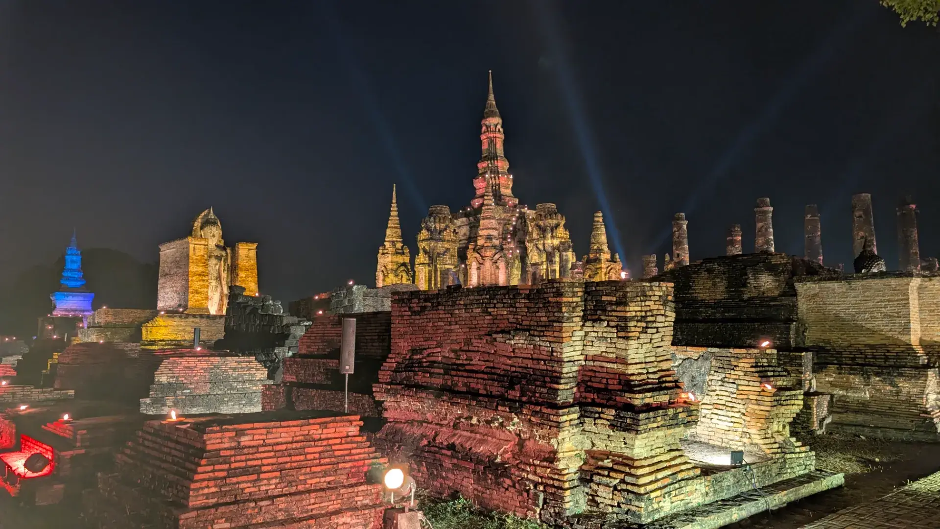夜のスコータイ歴史公園、ワット・マハータートの遺構。ライトアップされた仏塔と仏像から空に向かって数本のサーチライトが伸び、赤レンガの壁や基壇が幻想的に照らし出されている壮麗な景色。