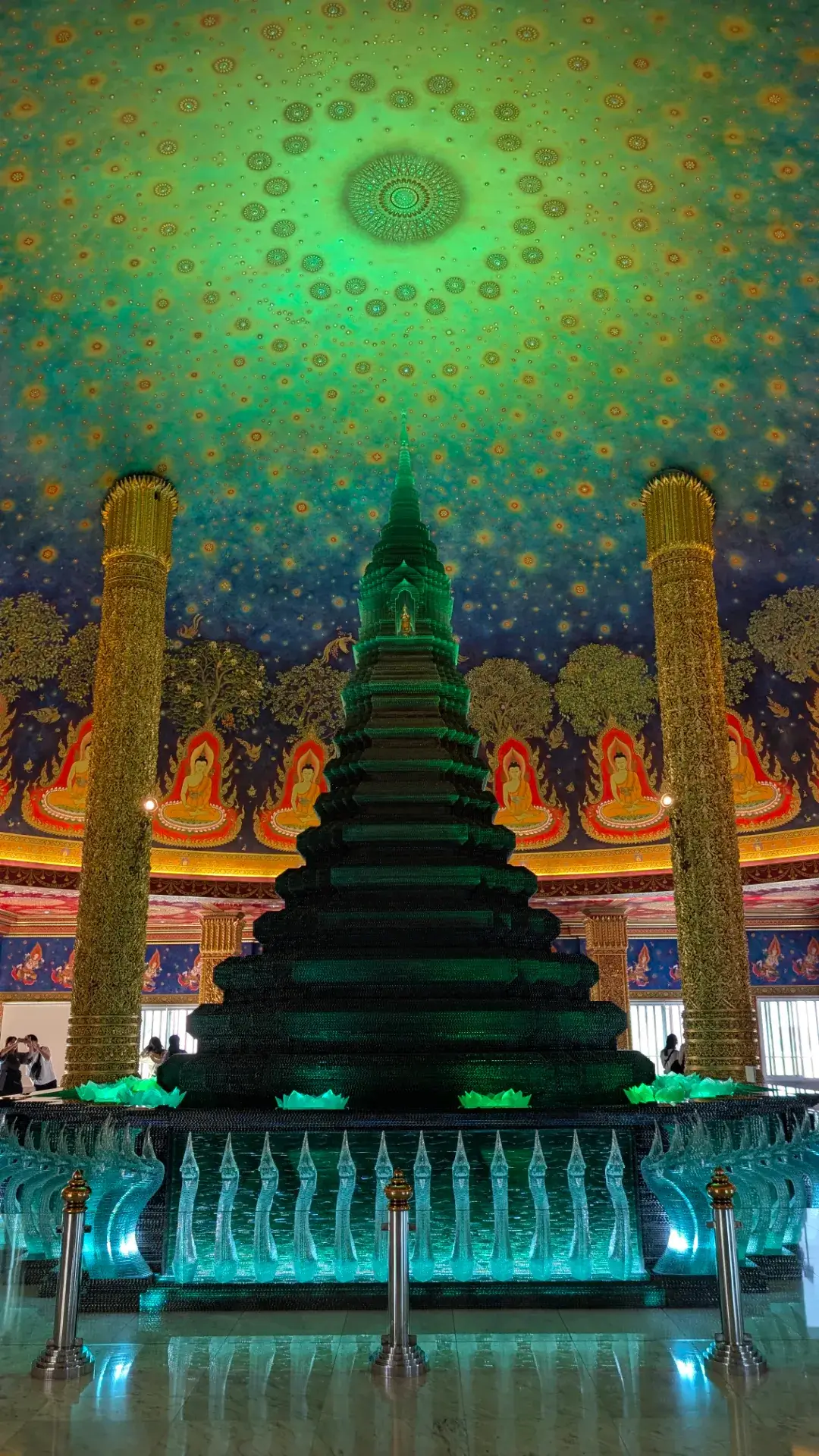 ワット・パクナムの大仏塔内、エメラルド色のガラス仏塔を正面から捉えた全景。仏塔の足元には、透き通るような緑色のガラスで精巧に作られた無数の龍（ナーガ）が鎮座し、守護神として仏塔を支えています。天井のドーム状に広がる宇宙的な仏教画と、黄金に輝く柱、そして幻想的な緑の光が織りなす空間は、まさにバンコクで最も美しい聖域の一つと言える圧倒的な光景