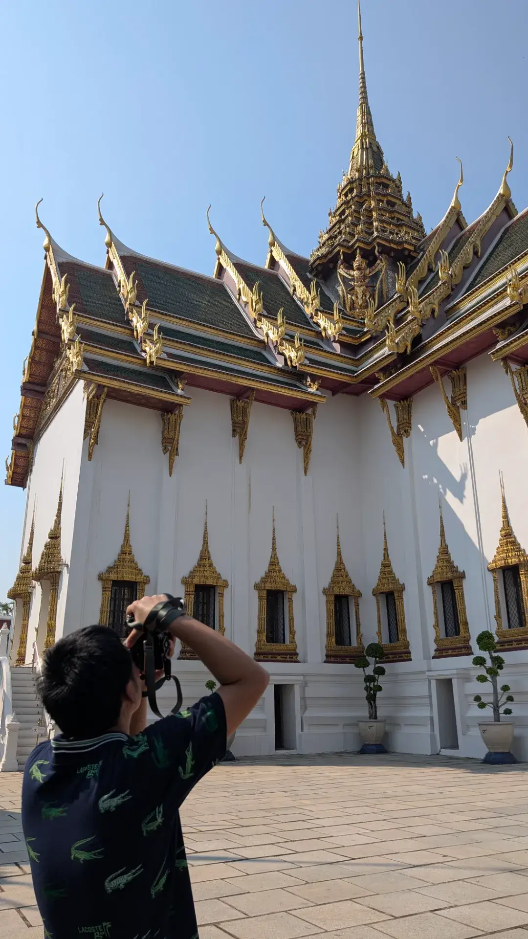 ワット・プラケオ（王室守護寺院）の敷地内に建つ、純白の壁と精巧な装飾の屋根が特徴的な「ドゥシット・マハ・プラサート宮殿」。ラーマ1世によって建てられたこの宮殿は、王室の儀式や葬儀などが行われる重要な場所です。青空に向かってそびえる尖塔と、伝統的なタイ様式の建築美を熱心に撮影する人々の姿が、歴史的聖域の静かな活気を伝えています。