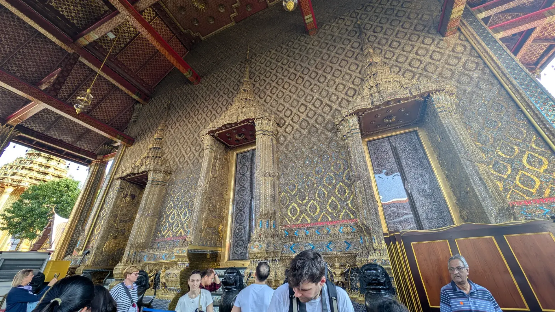 バンコクの王宮敷地内に位置する「ワット・プラケオ（Wat Phra Kaew）」の本堂外壁。金箔や色とりどりのガラス細工で埋め尽くされた壁面は、日光を浴びて神々しく輝いています。精巧な彫刻が施された窓や柱の装飾は、タイ最高峰の建築技術と芸術性の結晶であり、多くの参拝客や観光客がその圧倒的な美しさに足を止める、タイ観光の象徴的な光景です。