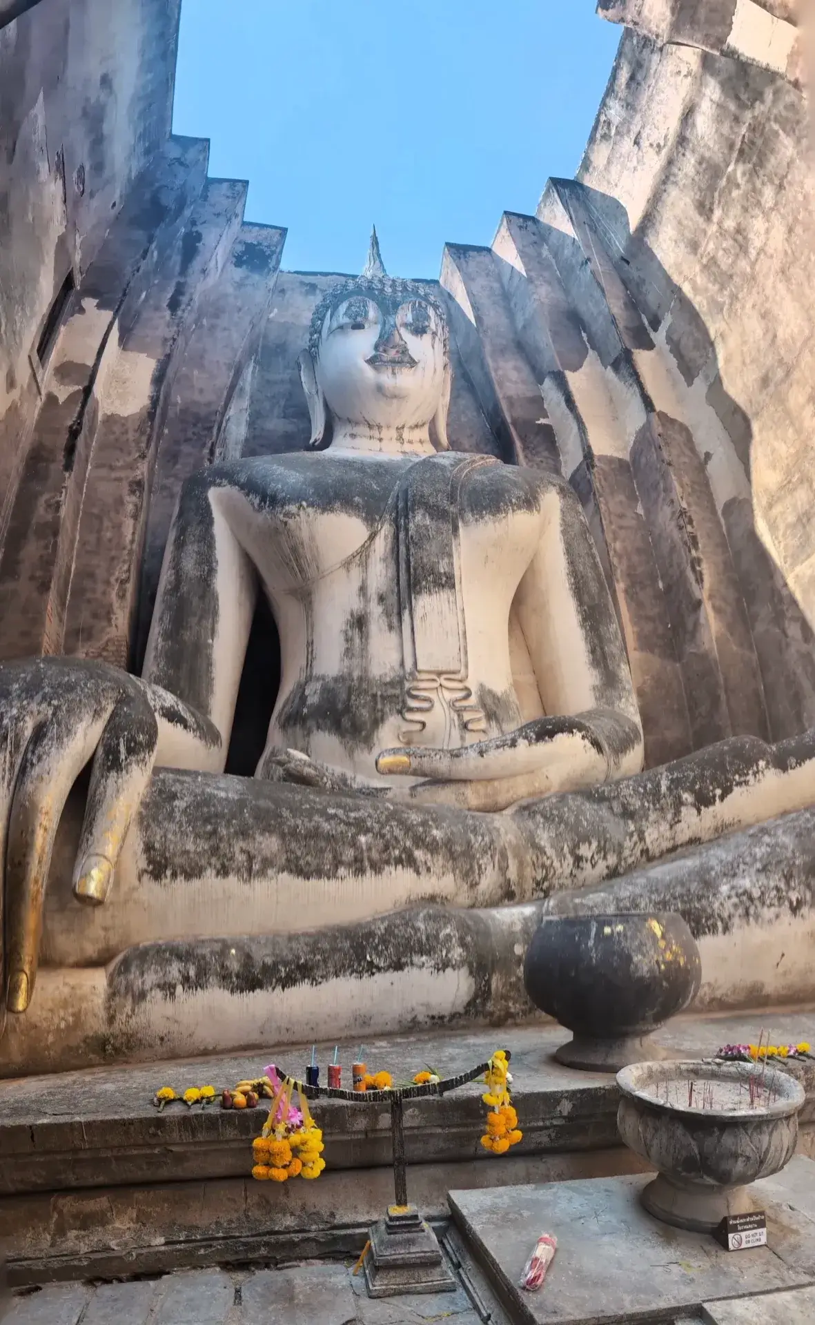 タイのスコータイ歴史公園にある「ワット・シーチュム（Wat Si Chum）」の内部に鎮座する、巨大なアチャナ仏の全景。屋根のないモンドップの空間に、高さ約15メートルの仏坐像が堂々と座しており、その穏やかな表情と、右手の指先に施された金箔の輝きが、訪れる者に深い慈悲と歴史の重みを感じさせる神秘的な光景です。