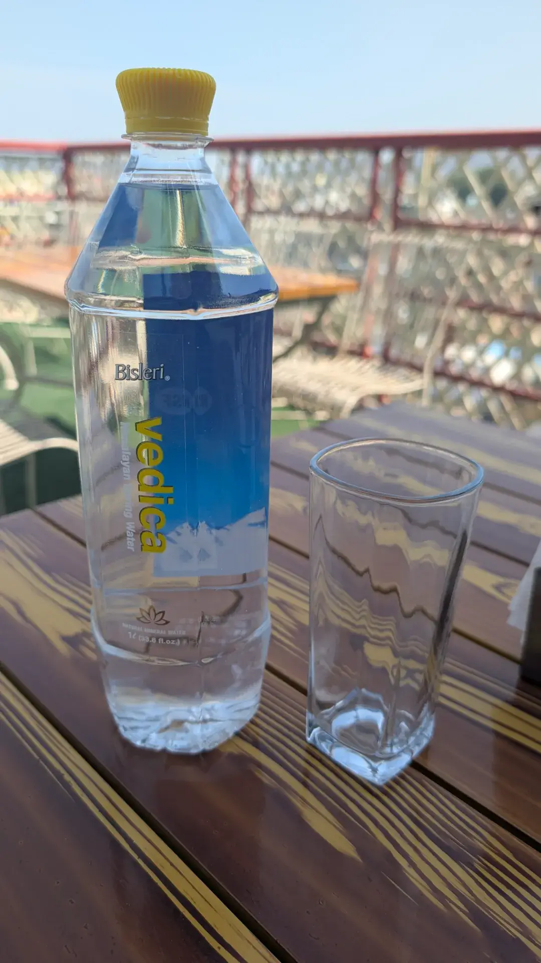 木製のテーブルの上に置かれた『ビスレリ・ヴェディカ（Bisleri Vedica）』のボトル入り飲料水と、その横にある空のグラス。背景にはテラス席の黒い柵と柔らかな光が差し込むカフェの空間が広がっている。