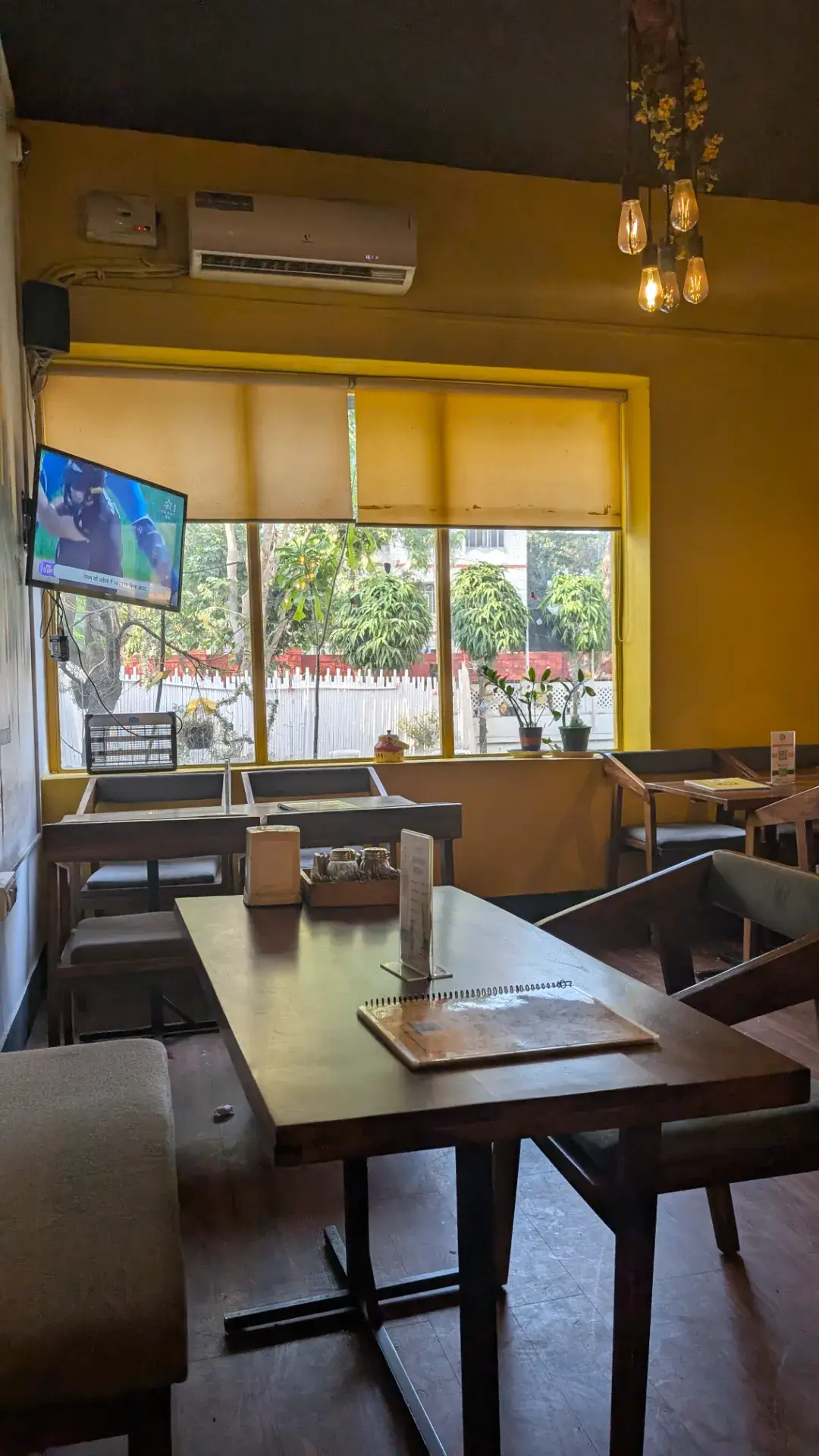 黄色い壁と木製家具が落ち着くカフェの店内。窓から外が見えるテーブル席とテレビ。