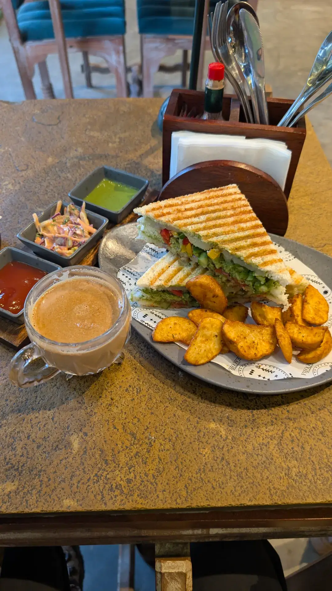 グリルされたサンドイッチとフライドポテトが盛り付けられたプレート、カップに入ったミルクティー（マサラチャイ）、そして3種類のソース（ケチャップ、コールスロー、緑色のチャツネ）が並ぶテーブル。