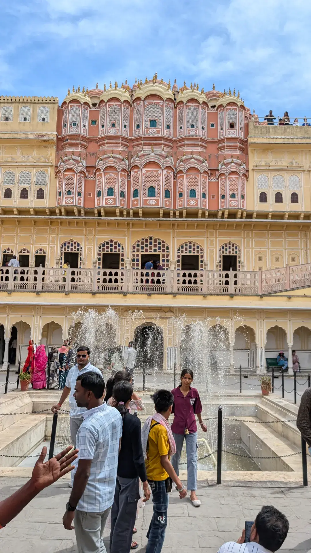 インド、ジャイプールのハワ・マハル（風の宮殿）の内部にある中庭。中央に涼しげな噴水があり、周囲には黄色とピンクの砂岩で彩られた繊細なアーチやバルコニーが広がる。多くの観光客が歴史的な建築美を楽しんでいる様子。