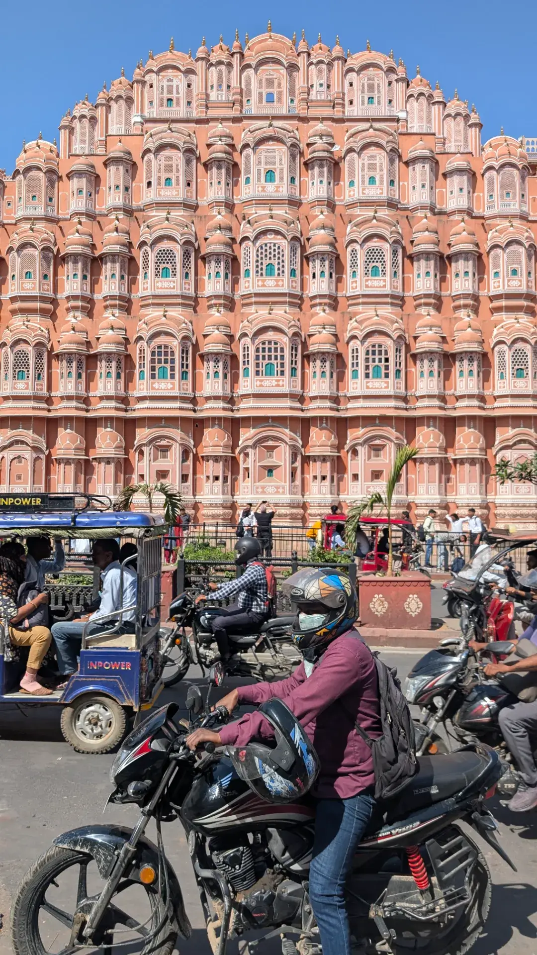 インド、ジャイプールの「ハワ・マハル（風の宮殿）」。953個の小窓が並ぶピンク色の砂岩でできた繊細なファサードを背景に、手前の道路を走るオートリキシャやバイク、行き交う人々が織りなすジャイプール特有の活気ある日常風景。