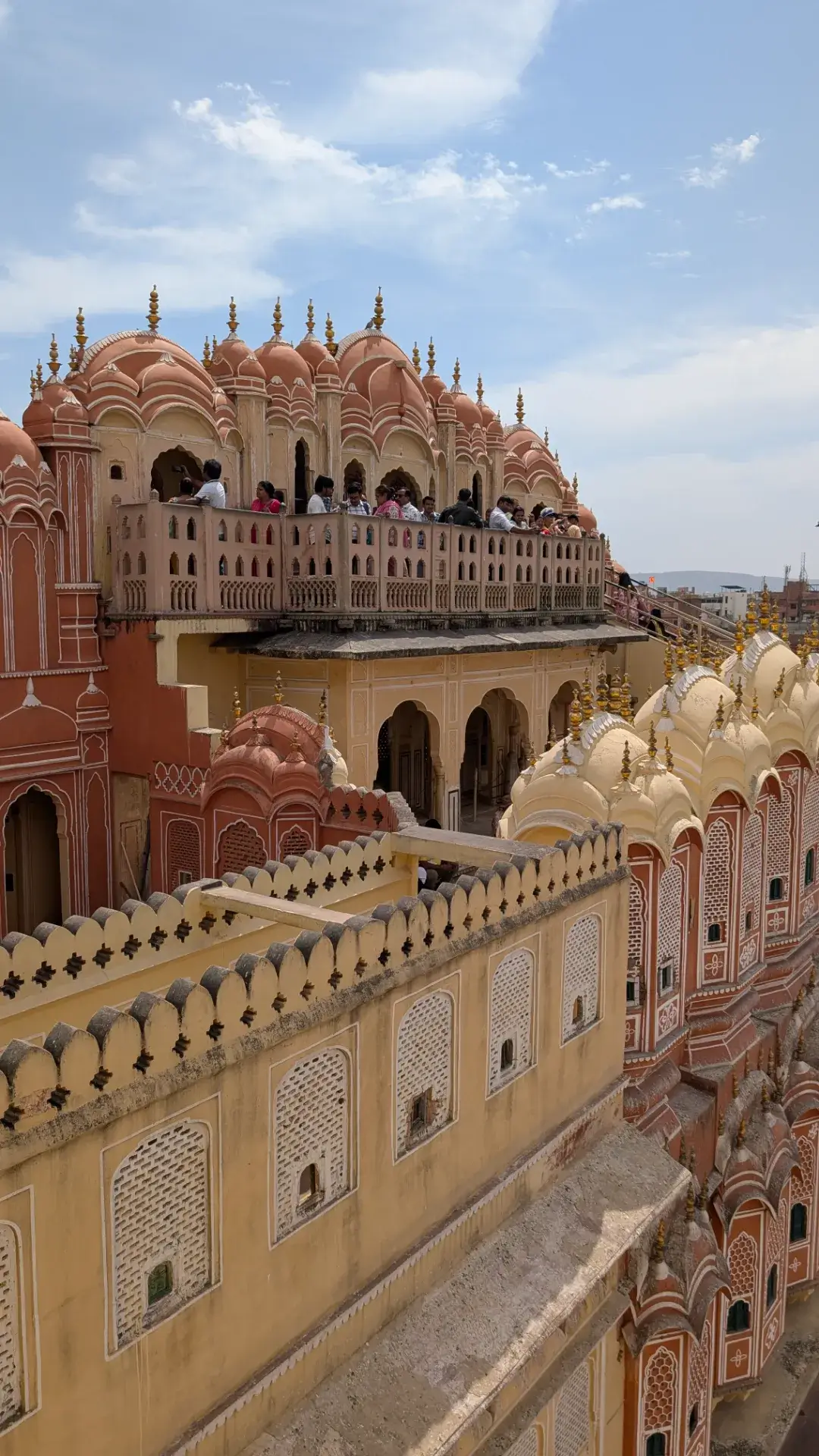 インド、ジャイプールのハワ・マハルの上層階。ピンク色と黄色の砂岩で造られた多層構造のバルコニーに観光客が並び、手前には精巧な彫刻が施された城壁とドーム状の屋根（チャトリ）が連なる、立体的な建築風景。