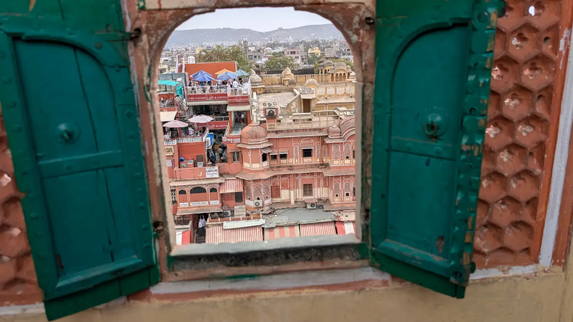 ジャイプールのハワ・マハル（風の宮殿）にある、緑色の小さな開き戸がついた窓から見た街の風景。正面にはオレンジやピンク色の建物が並び、屋上テラスのカフェや遠くの山々まで見渡せる、歴史と現代が交差する景色。
