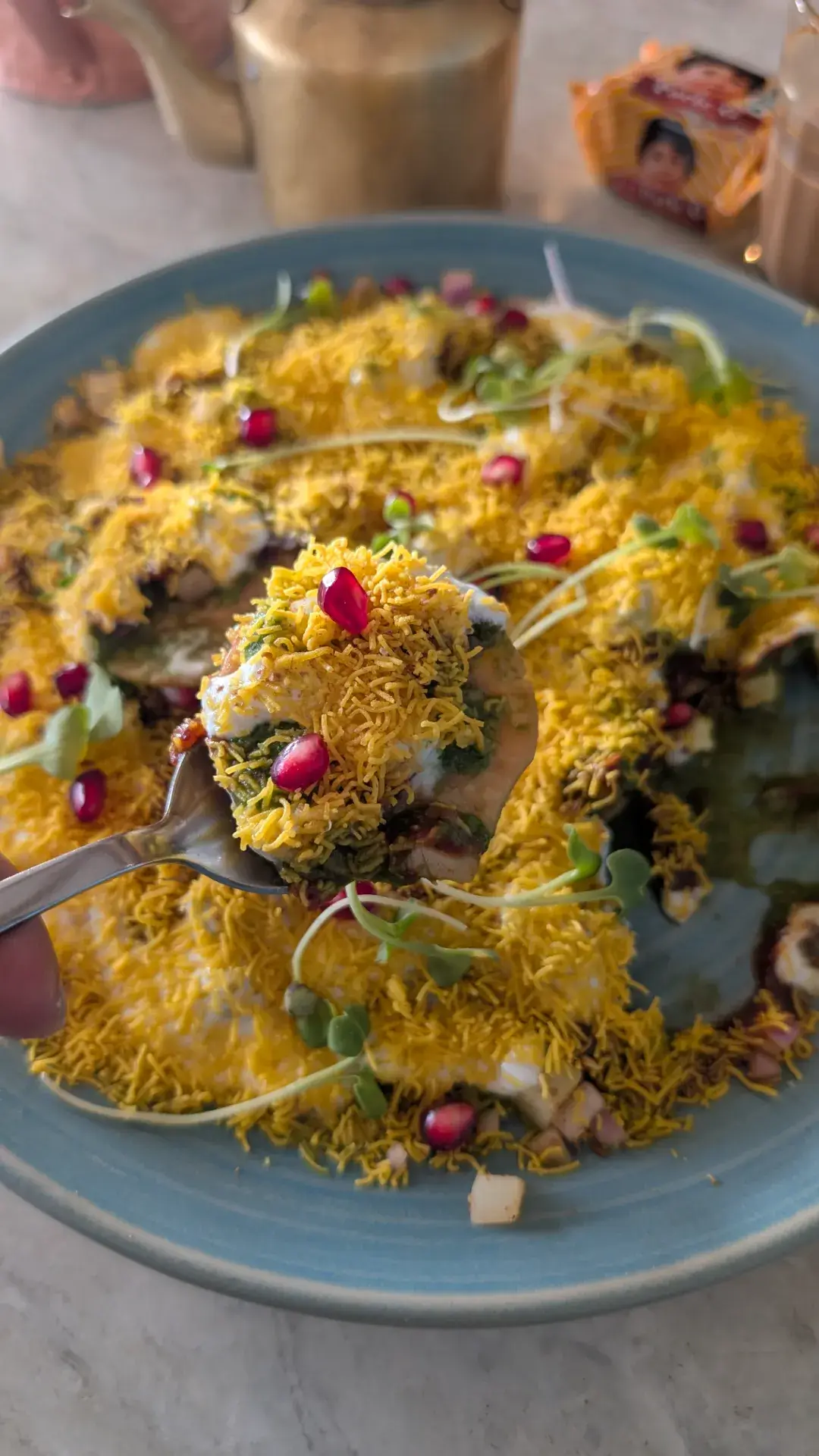 ジャイプールのカフェでパプリ・チャートを食べる様子。スプーンの上に、ソースの染みたパプリ（揚げせんべい）、たっぷりのセブ（揚げ麺）、緑のミントチャツネ、白のヨーグルト、そして宝石のように輝く赤いザクロが完璧な層となって乗っている。