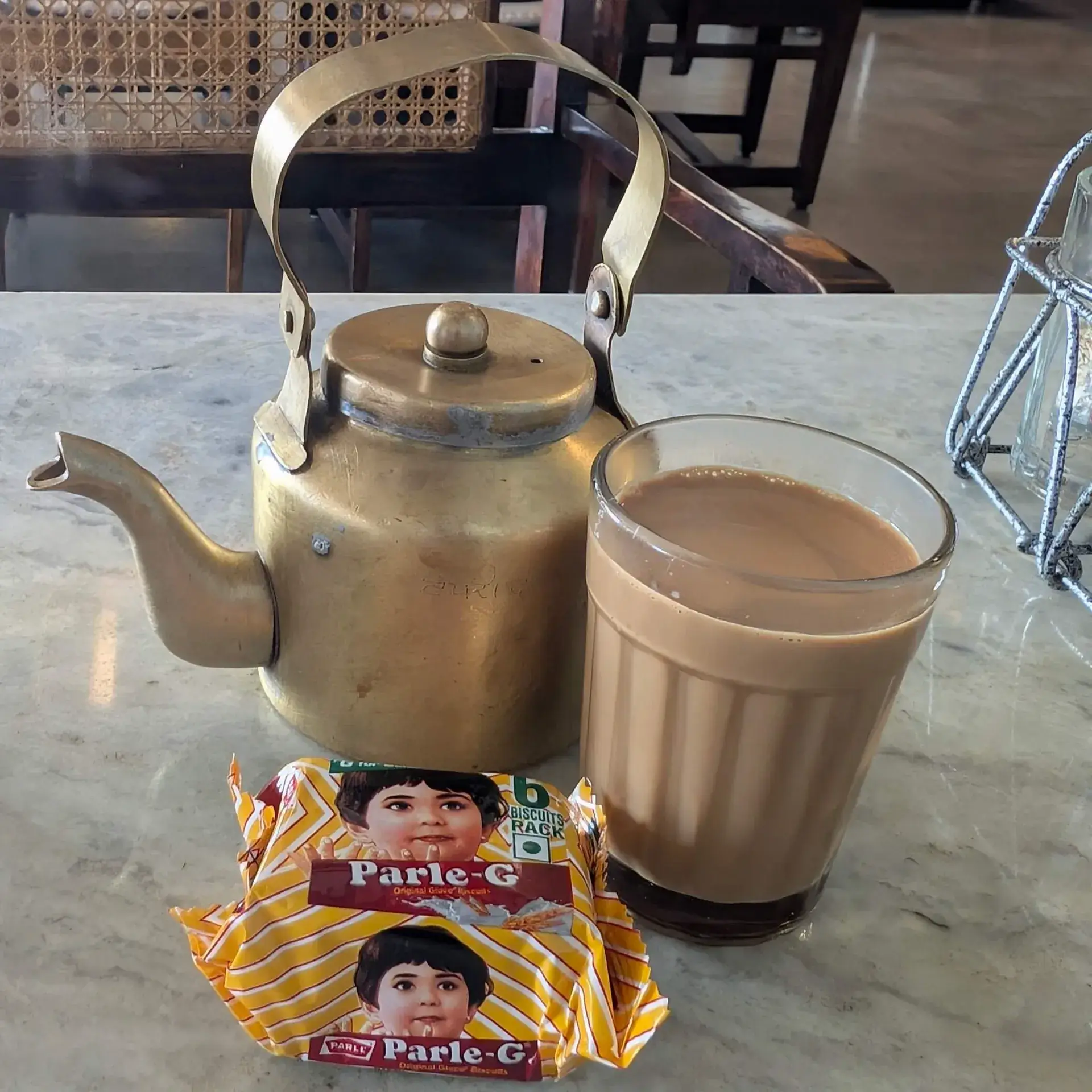 インド、ジャイプールのカフェで提供された伝統的なチャイ。使い込まれた真鍮のケトル、グラスに注がれたミルクティー（チャイ）、そして手前にはインドで最も有名な「Parle-G（パール・ジー）」ビスケットのパックが置かれた、大理石のテーブルの風景。