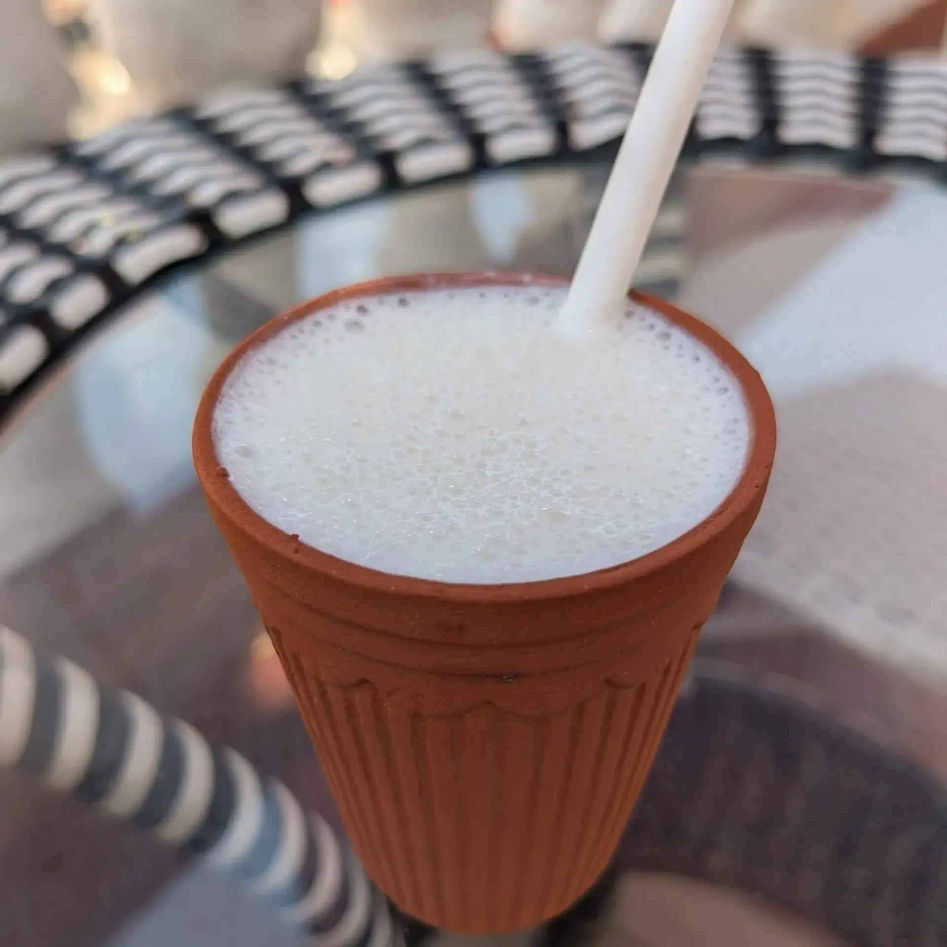 インド、ジャイプールのカフェで提供された伝統的なラッシー。使い捨ての素焼きのカップ（クッラル）に注がれ、表面が白く泡立った濃厚なヨーグルトドリンク。背景にはカフェのテーブルの質感がボケて写っている。