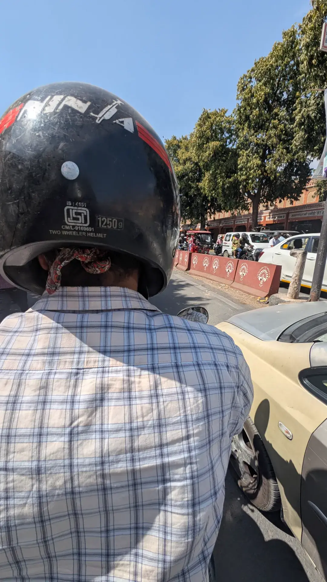 ジャイプールの街を走るUber Moto（バイクタクシー）の後部座席からの視点。チェック柄のシャツを着て黒い「NINJA」ヘルメットを被った運転手の背中越しに、ピンク色の建物が並ぶジャイプール特有の街並みと、並走するリキシャや自動車の混雑が見える。
