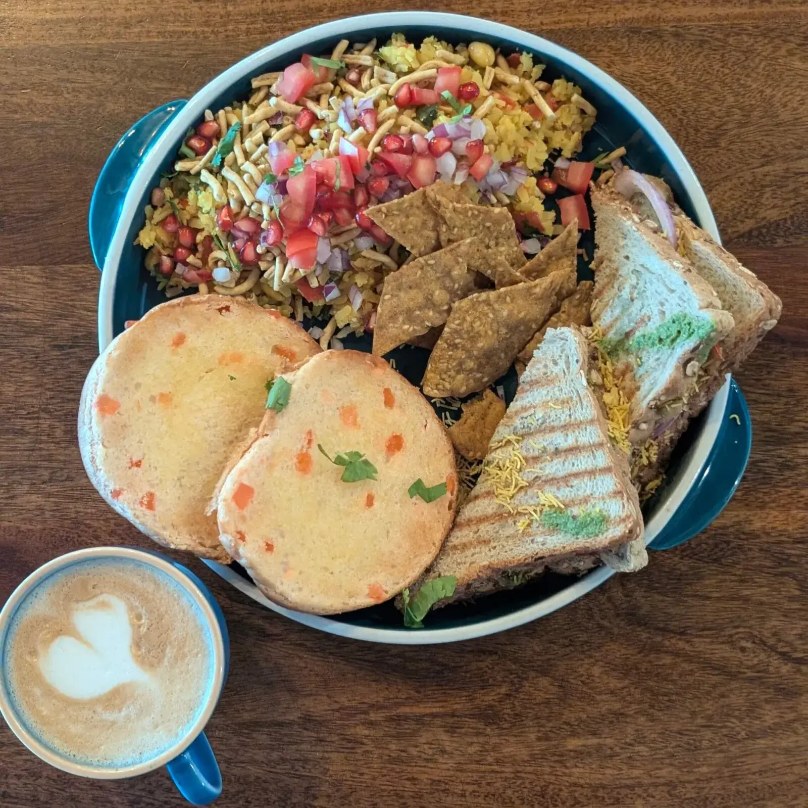 大きな皿に盛り付けられたインドの朝食セット。ポハ（平らな米の炒め物）にトマトやザクロがトッピングされたもの、グリルサンドイッチ、パンの付け合わせ、そしてハート型のアートが描かれたカフェラテが並ぶ食卓。