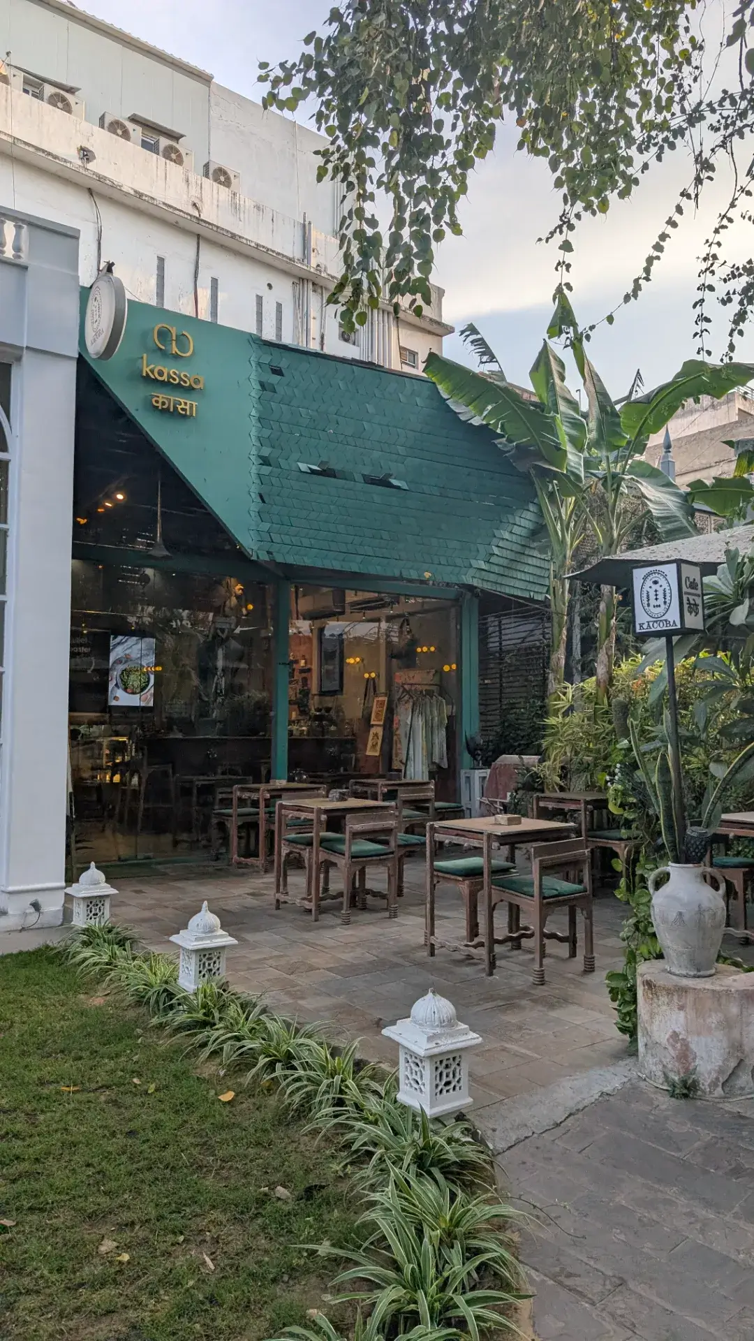 緑色の屋根が特徴的な『kassa（カサ）』というカフェの外観。テラス席には木製のテーブルと椅子が並び、周囲は観葉植物や白いガーデンライトで装飾されている。