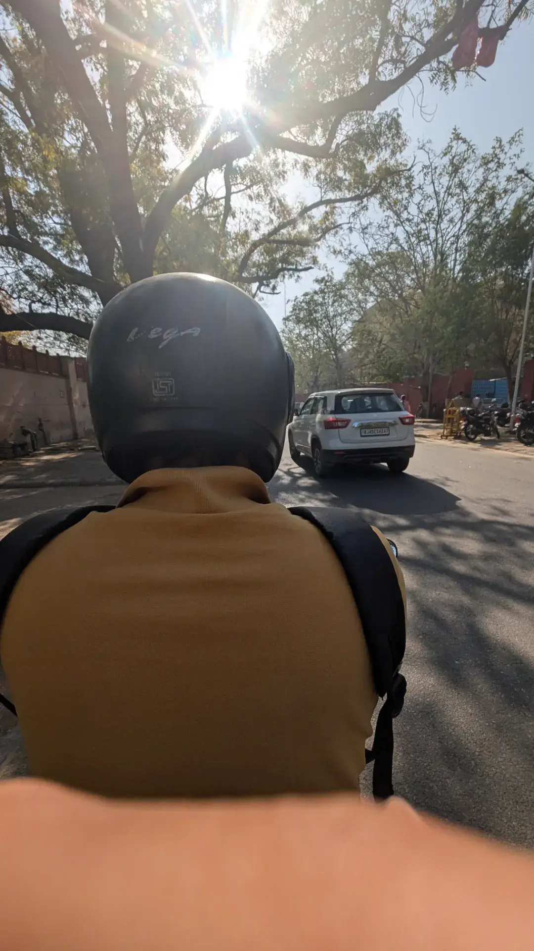 バイクの後部座席から見た、街中の道路を走る風景。眩しい太陽の光が木々の間から差し込み、前方を走る白いSUVと周辺の街並みが写っている。