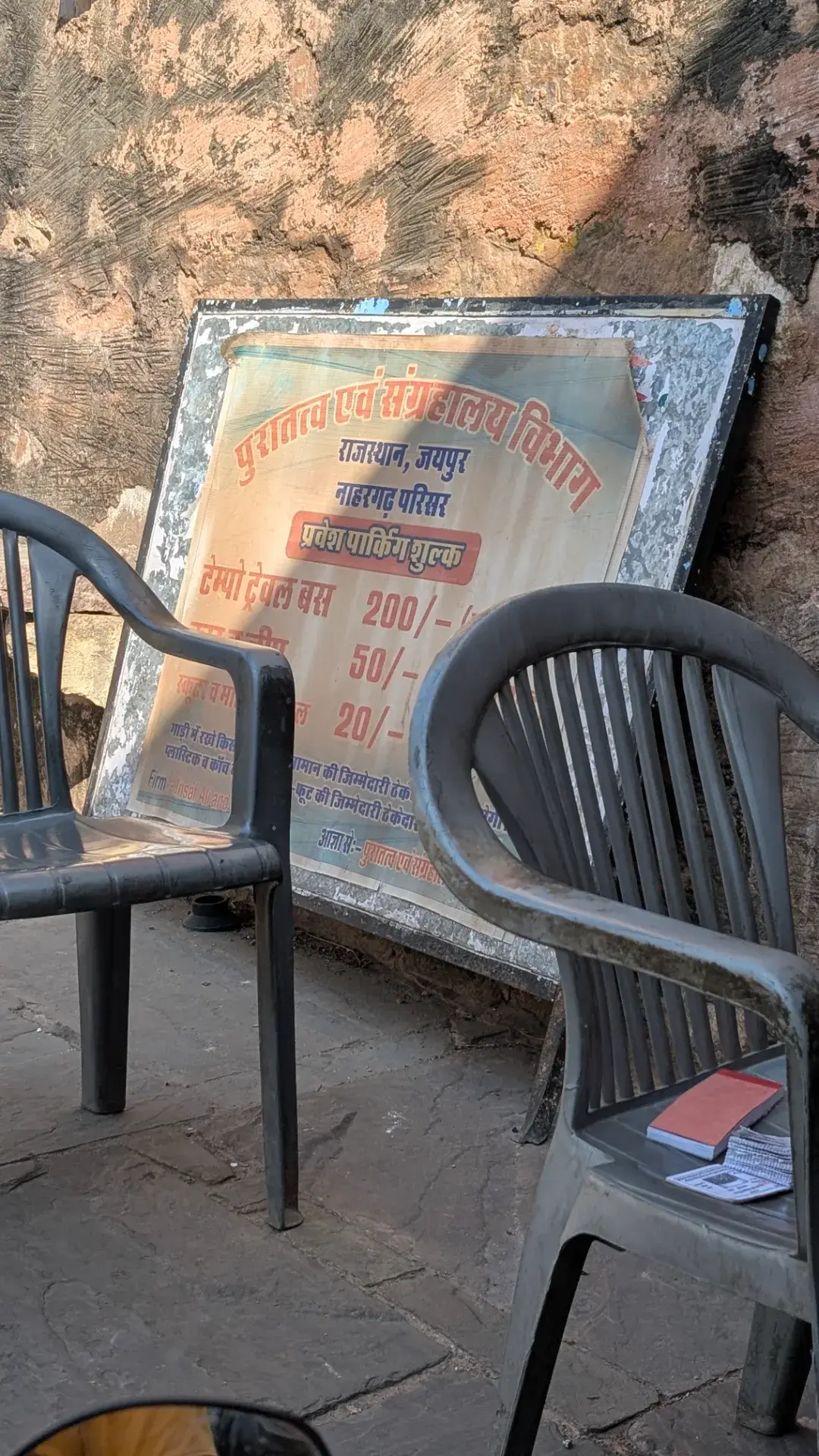 インドの歴史的遺跡の入り口に設置された駐車料金表の看板。石壁を背景に、手前には休憩用のプラスチック製の椅子が置かれ、右側の椅子の上にはメモ帳と小銭が乗っている。