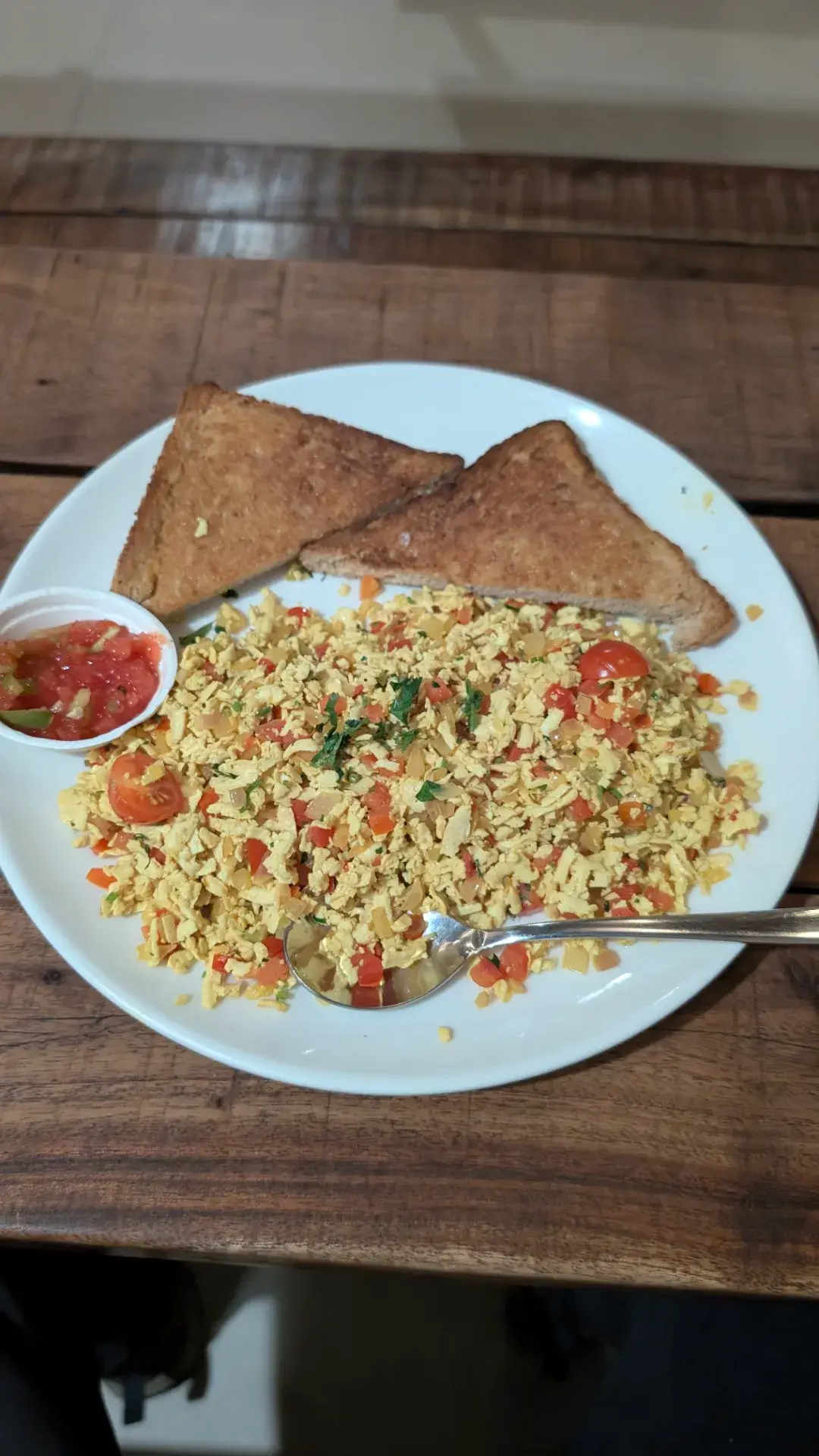 白い皿に盛り付けられた、トマトや玉ねぎなどの野菜を混ぜ込んだスクランブルエッグと、三角形にカットされたトースト2枚、小さなカップに入ったサルサソース。