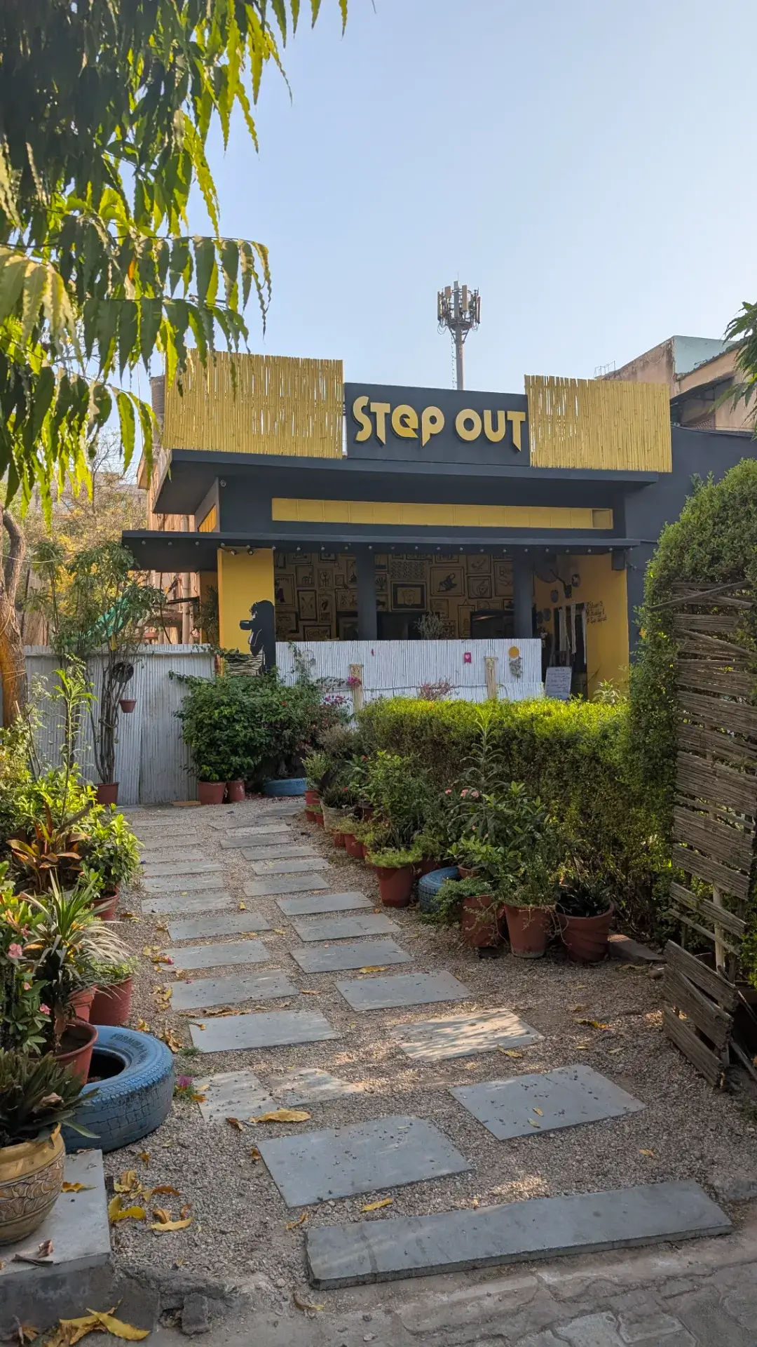 『STEP OUT』という黄色い看板が掲げられたカフェの外観。石畳の小道の両脇に観葉植物が並び、黄色と黒を基調としたモダンで開放的な入り口が特徴。