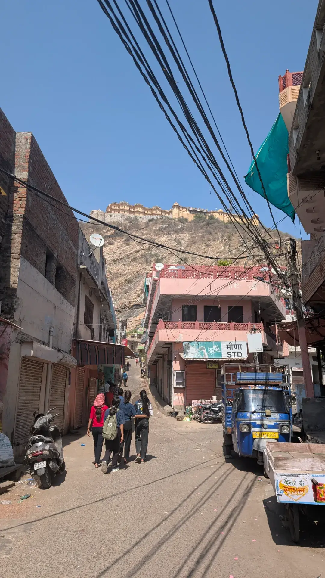 アンベール城を遠くに見上げるジャイプールの街並み。人々が歩く細い通りと、手前のオート三輪やバイクの風景。