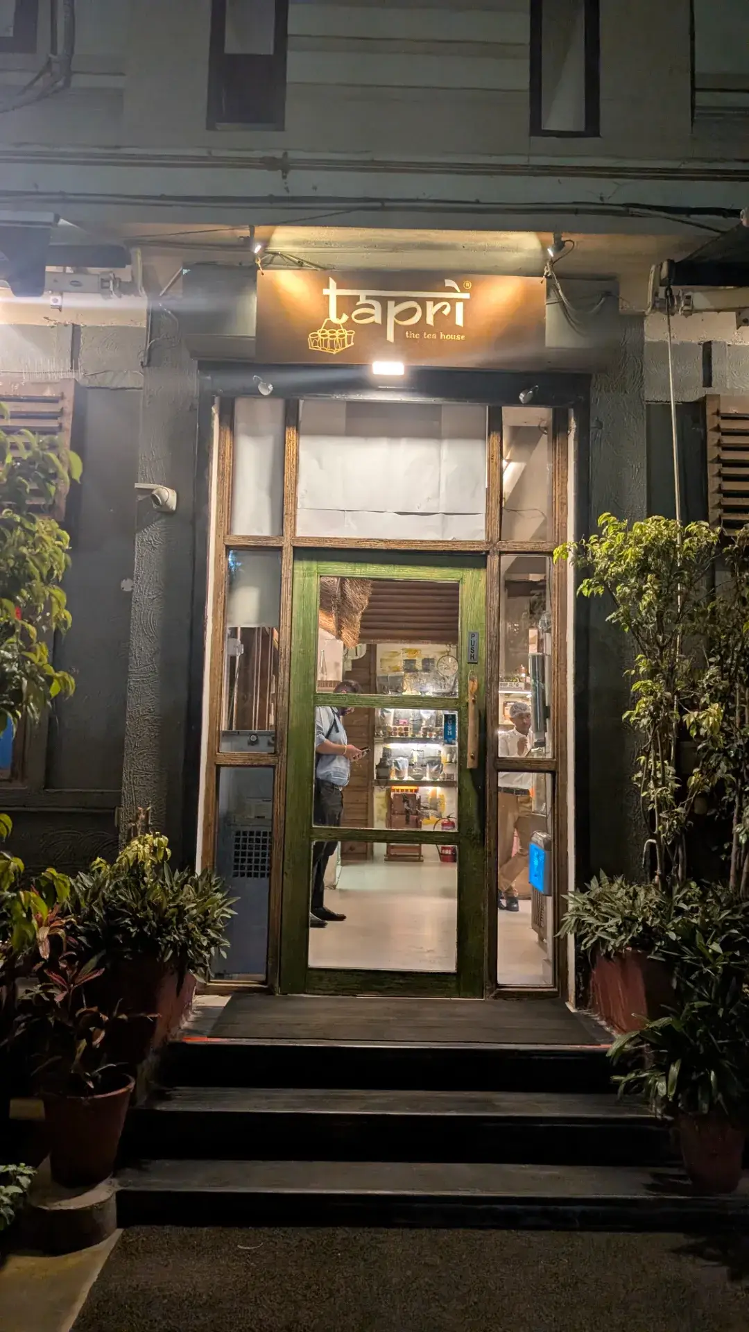 夜間に撮影された、暖色系のライトに照らされた『tapri the tea house』という看板があるカフェの入り口。緑色の枠のガラスドアから、店内の様子がうかがえる。