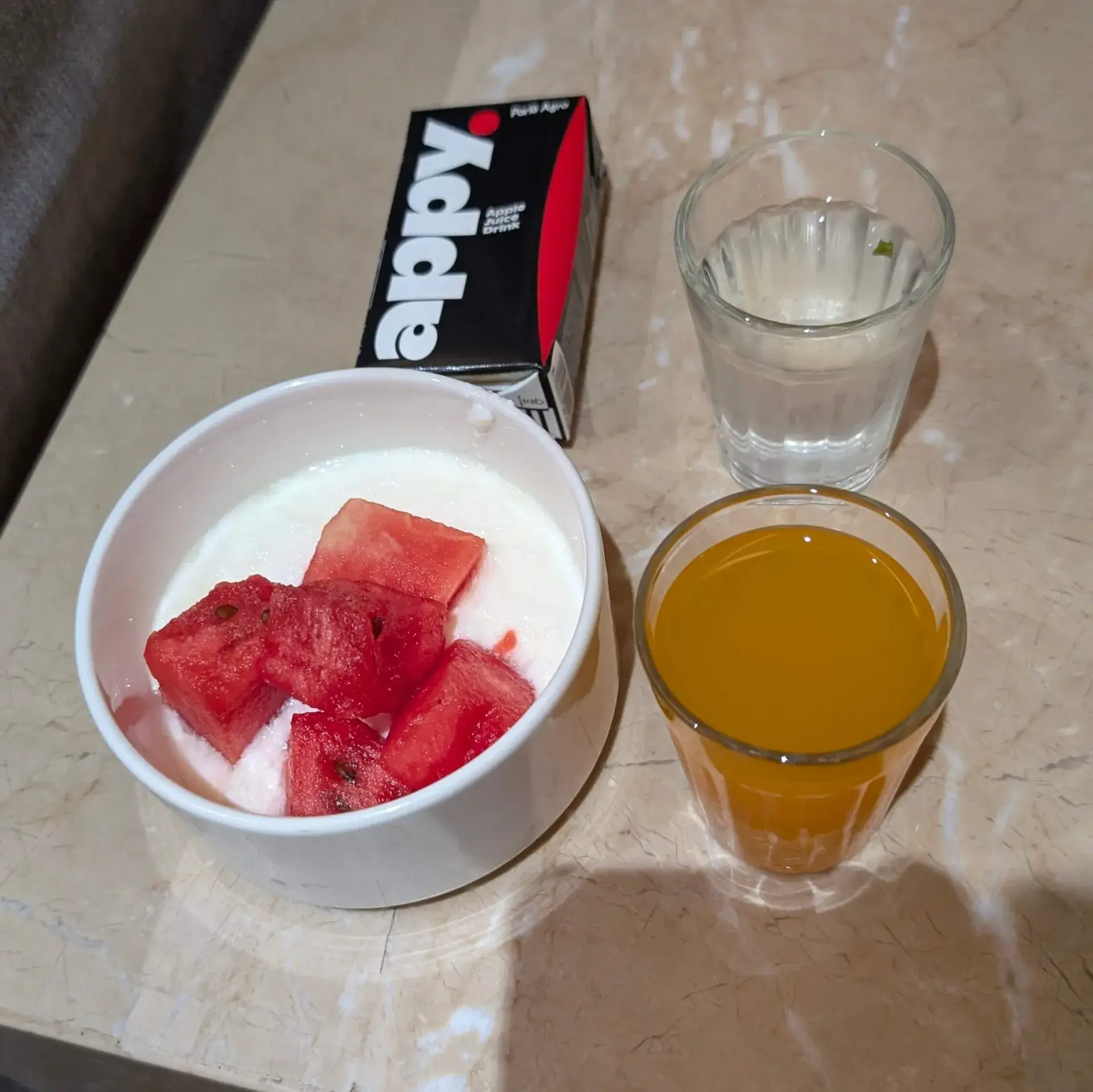 ボウルに入ったスイカのヨーグルト和えと、紙パックのアップルジュース、水、オレンジジュースの朝食セット。